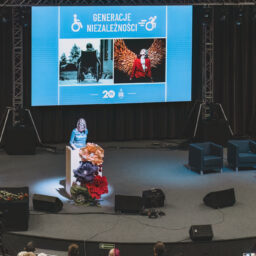 Widok na ciemną scenę. Po lewej stronie za mównicą stoi elegancka kobieta. Za nią na ekranie na niebieskim tle na górze biały napis: Generacje Niezależności; po obu stronach ikony osób poruszających się na wózkach. Poniżej po lewej czarno-białe zdjęcie dziecka siedzącego tyłem na dużym, pokojowym wózku. Po prawej kolorowe zdjęcie pięknej młodej dziewczyny patrzącej w górę, siedzi na wózku, za nią złote skrzydła. Na dole pośrodku białe logo Fundacji PODAJ DALEJ i logo wpisane w liczbę 20 na niebieskim pasku.