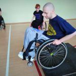 Na sali gimnastycznej młody chłopak siedzący na wózku inwalidzkim zjeżdża z niskiej przeszkody imitującej krawężnik. W tle dwie inne osoby na wózku przyglądają się tej czynności.