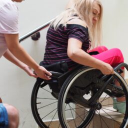 Kobieta na wózku zjeżdża tyłem po schodach z pomocą asystenta