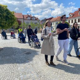 Na zdjęciu grupa osób na brukowanym rynku. Jest słoneczny dzień. Część osób porusza się na wózkach inwalidzkich, a część idzie pieszo. Na pierwszym planie kobieta w jasnym płaszczu idzie pod rękę z młodym mężczyzną w sportowym stroju. Obok nich młoda kobieta prowadzi starszą panią na wózku. W tle widać kolejne osoby, zarówno na wózkach, jak i chodzące pieszo, a także budynki otaczające rynek.