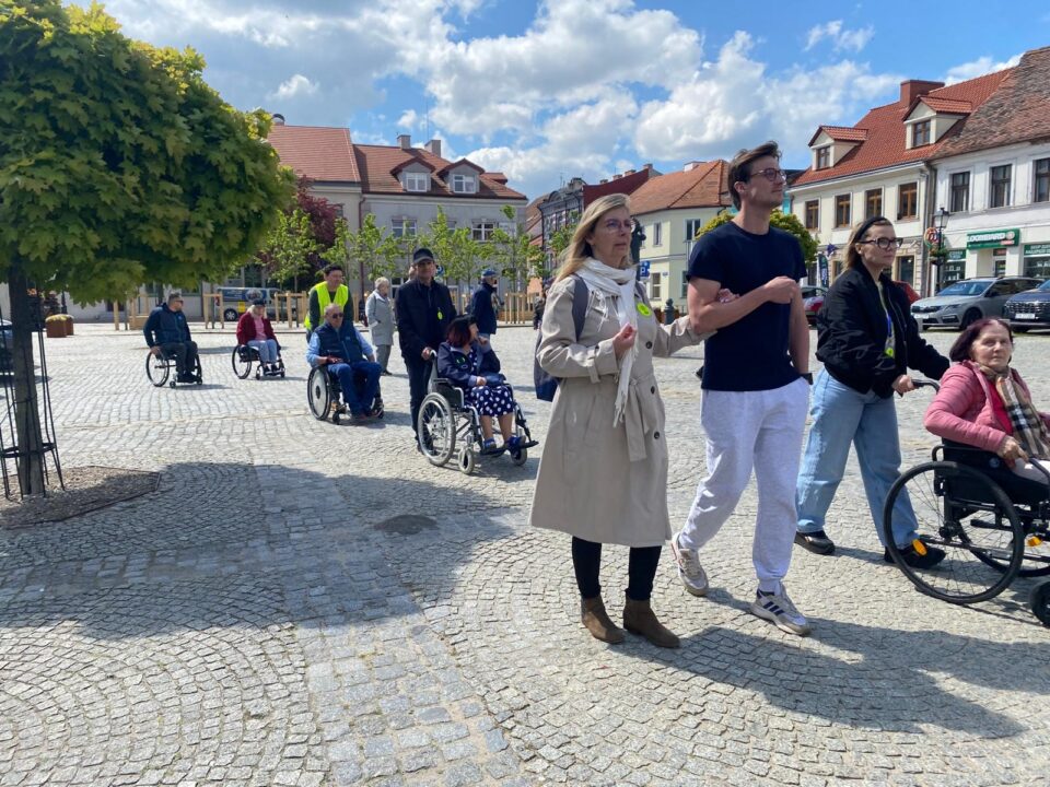 Na zdjęciu grupa osób na brukowanym rynku. Jest słoneczny dzień. Część osób porusza się na wózkach inwalidzkich, a część idzie pieszo. Na pierwszym planie kobieta w jasnym płaszczu idzie pod rękę z młodym mężczyzną w sportowym stroju. Obok nich młoda kobieta prowadzi starszą panią na wózku. W tle widać kolejne osoby, zarówno na wózkach, jak i chodzące pieszo, a także budynki otaczające rynek.