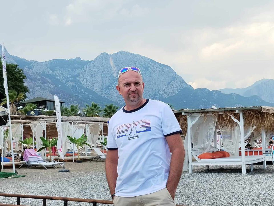 Mężczyzna ubrany w biały t-shirt uśmiecha się do zdjęcia stojąc na tle miejsca wypoczynkowego na plaży.