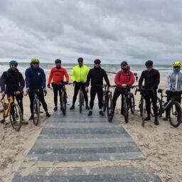 Grupa 8 mężczyzn - rowerzystów na nadmorskiej plaży. Pozują do zdjęcia z rowerami, ubrani w stroje przeciwdeszczowe. Pochmurno.