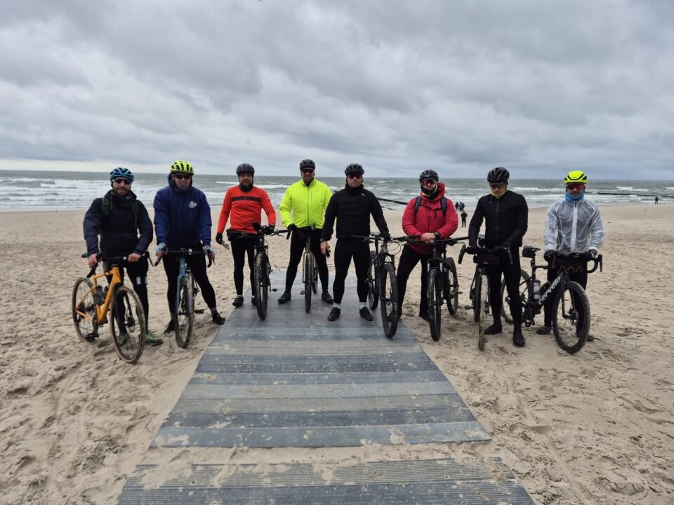 Grupa 8 mężczyzn - rowerzystów na nadmorskiej plaży. Pozują do zdjęcia z rowerami, ubrani w stroje przeciwdeszczowe. Pochmurno.