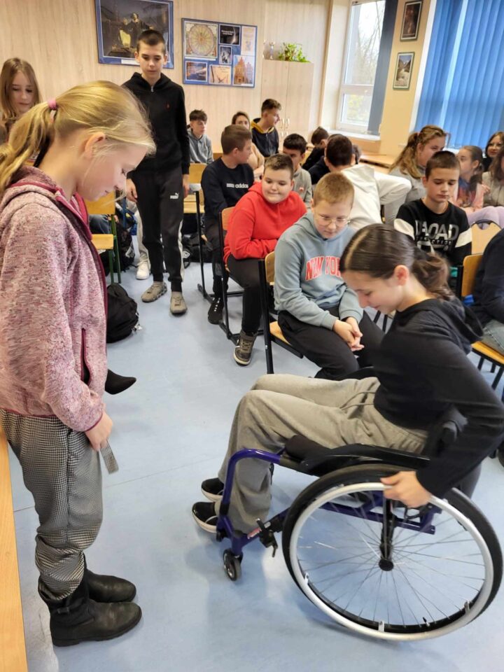 Jedna dziewczynka siedzi na wózku, druga stoi przed nią i ją obserwuje. W tle cała klasa siedzi na krzesłach i patrzy na dziewczynki