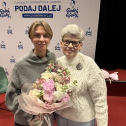 Dwie kobiety w średnim wieku, uśmiechnięte, pozują do zdjęcia. Jedna z nich trzyma kwiaty. Za nimi ścianka Fundacji PODAJ DALEJ.