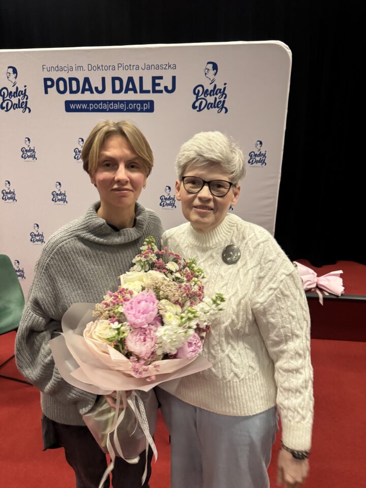 Dwie kobiety w średnim wieku, uśmiechnięte, pozują do zdjęcia. Jedna z nich trzyma kwiaty. Za nimi ścianka Fundacji PODAJ DALEJ.