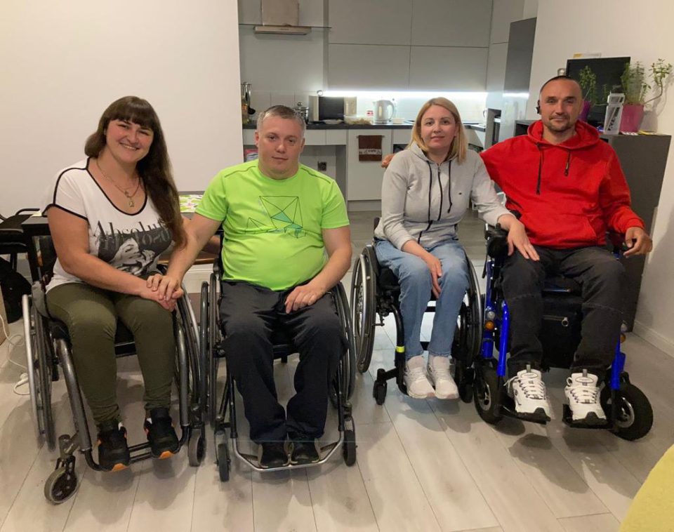 Na zdjęciu cztery dorosłe osoby na wózkach inwalidzkich - od lewej ciemnowłosa kobieta trzyma za rękę mężczyznę, dalej kobieta (blondynka) i mężczyzna w czerwonej bluzie.