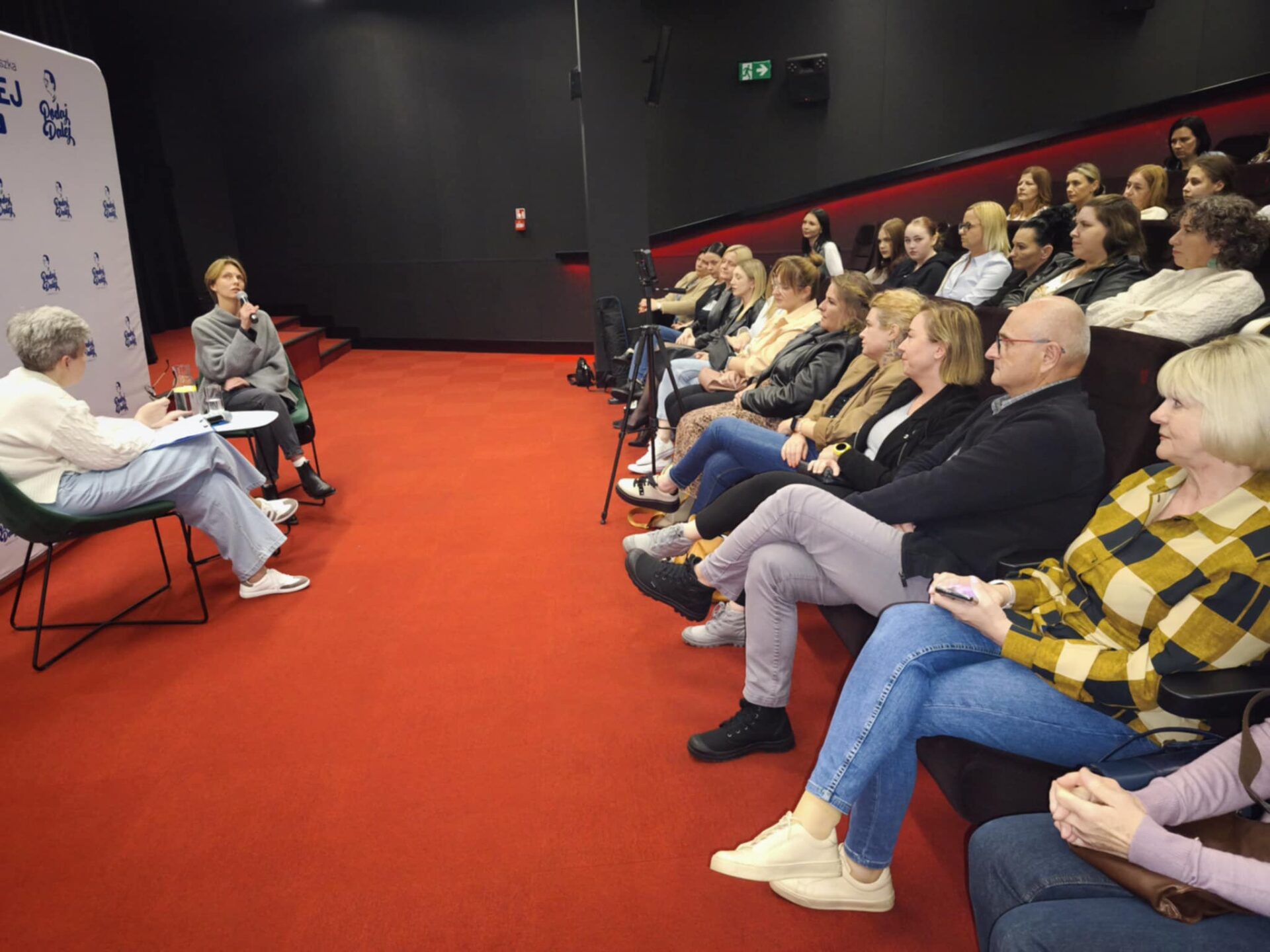 Sala widowiskowa, czerwona podłoga. Po prawej siedzą przy stoliku dwie kobiety w średnim wieku, jedna mówi do mikrofonu. Po prawej widać pełną widownię.