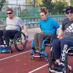 Dwóch młodych mężczyzn na wózkach, a między nimi młoda kobieta na wózku. Jadą po bieżni - każdy jest na swoim torze. Ubrani w lekkie stroje, pogoda słoneczna, panowie mają ciemne okulary. W tle widać krzesełka na widowni z zielonymi siedziskami.