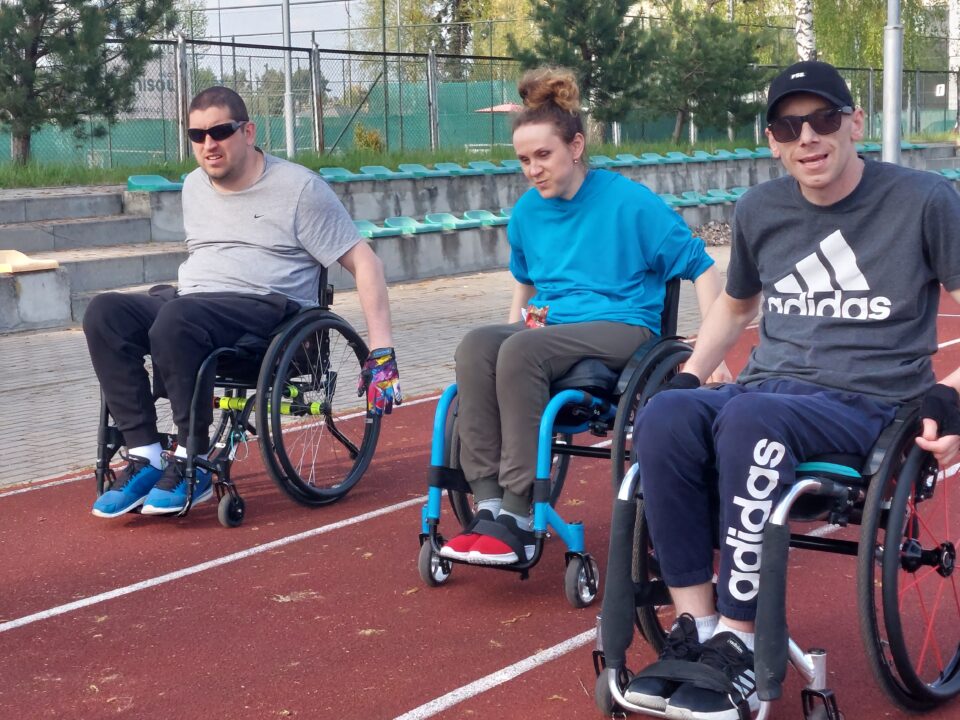 Dwóch młodych mężczyzn na wózkach, a między nimi młoda kobieta na wózku. Jadą po bieżni - każdy jest na swoim torze. Ubrani w lekkie stroje, pogoda słoneczna, panowie mają ciemne okulary. W tle widać krzesełka na widowni z zielonymi siedziskami.