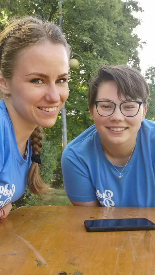 Zbliżenie na dwie uśmiechnięte młode kobiety w niebieskich koszulkach przy drewnianym stole na świeżym powietrzu. Jedna z nich ma warkocz, druga krótkie ciemne włosy i okulary. W tle zieleń