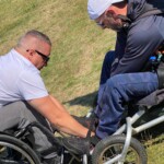 Po lewej mężczyzna na wózku inwalidzkim schyla się i poprawia coś przy nogach osobie, która siedzi na wózku do latania na paralotni