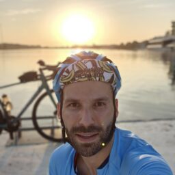 Zdjęcie przedstawia młodego mężczyznę, uśmiechnięty patrzy w obiektyw. Ma ciemne krótkie włosy i zarost, na głowie kolorowy kask rowerowy. Ubrany jest w jasną koszulkę rowerową - z nadrukami na ramionach i przodzie. Za sylwetką bohatera podcastu widoczna część ustawionego bokiem roweru. W tle jezioro, słabo widoczne fragmenty budynku po prawej stronie, w dalekim tle zachodzące słońce i zarysy drzew.